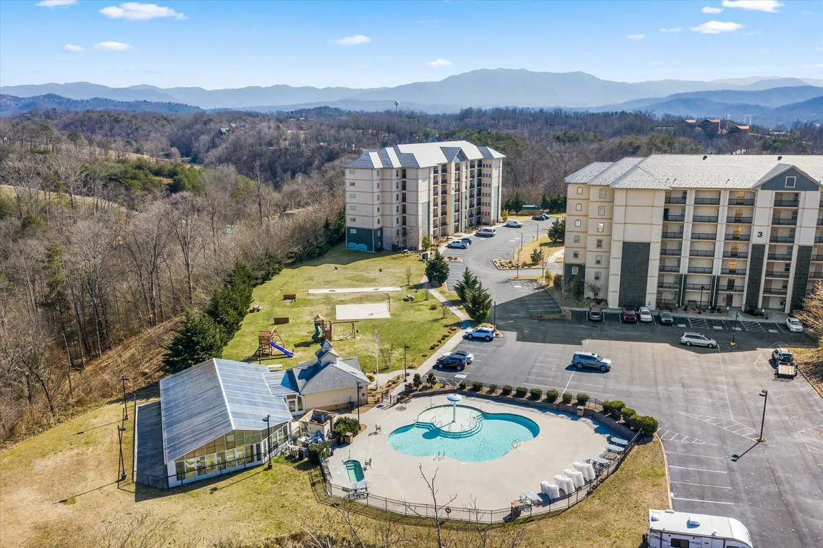 An exterior view featuring a pool area with clear blue water, surrounded by multi-story resort buildings. In the background, there is a playground and scenic mountain views, creating a vibrant family-friendly resort atmosphere. Lounge chairs and green spaces invite relaxation and play.