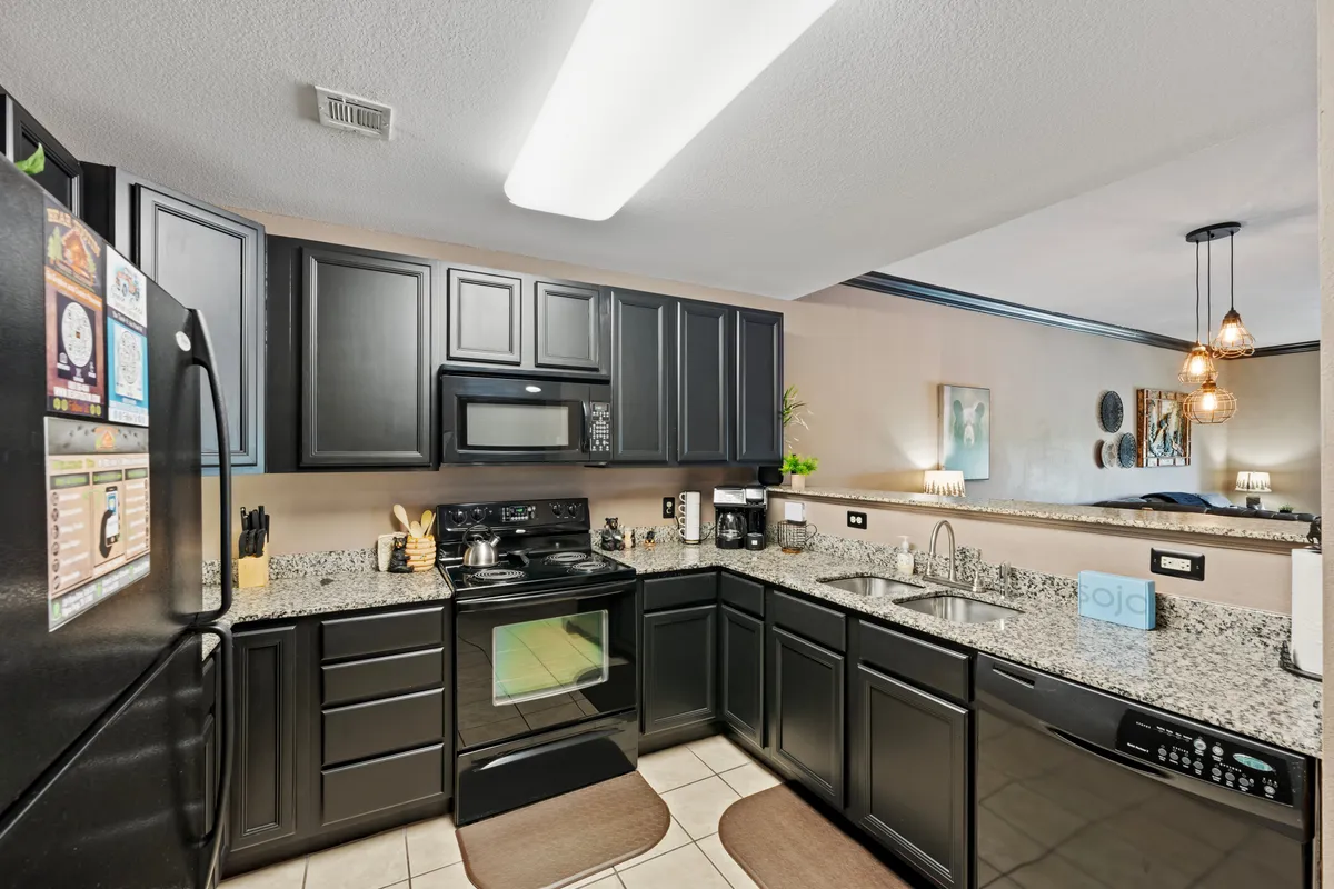 A view of a modern kitchen featuring black appliances, granite countertops, dark cabinetry, and a bar area illuminated by pendant lights. The kitchen provides ample space for meal preparation and socializing, with a warm color palette and contemporary finishes.