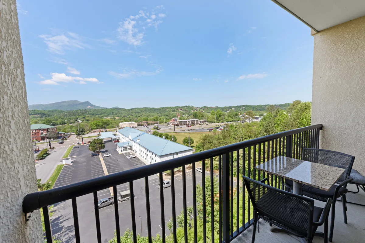 A serene, furnished balcony with comfortable seating set against a backdrop of sweeping mountain views and lush surrounding landscape. The scene evokes relaxation, with the sky and scenery visible beyond the railings.
