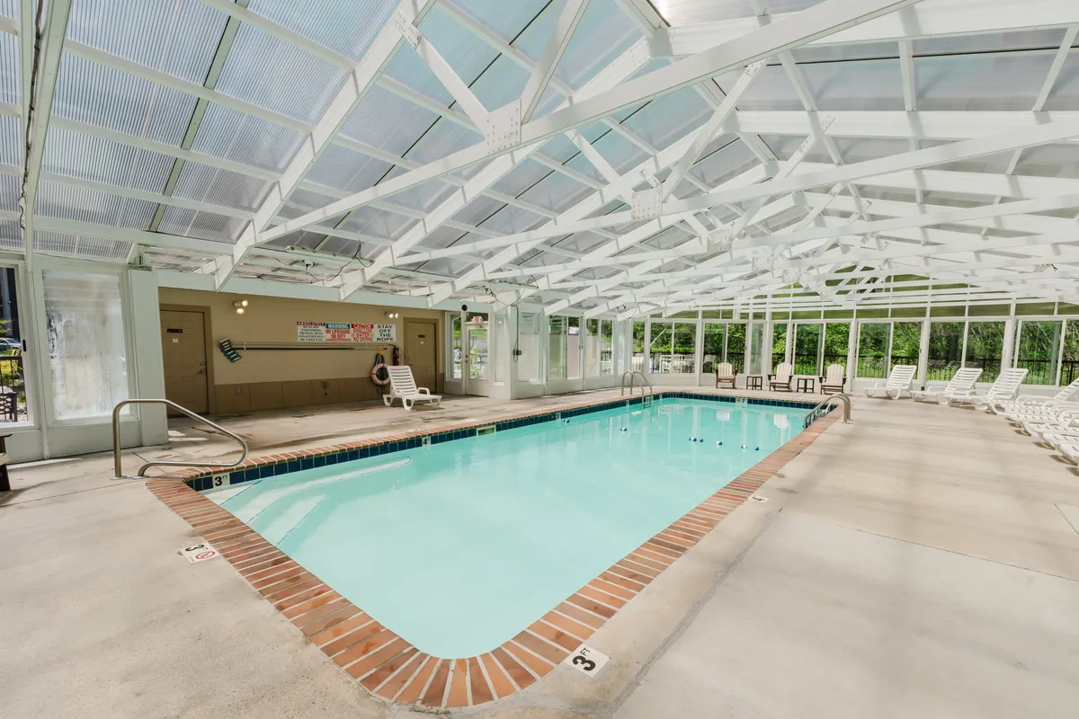 A bright and airy indoor swimming pool area with a large glass ceiling streaming in sunlight, lounge chairs for relaxation, and a tranquil mood. The space invites guests to unwind and enjoy the resort's amenities in comfort and style.