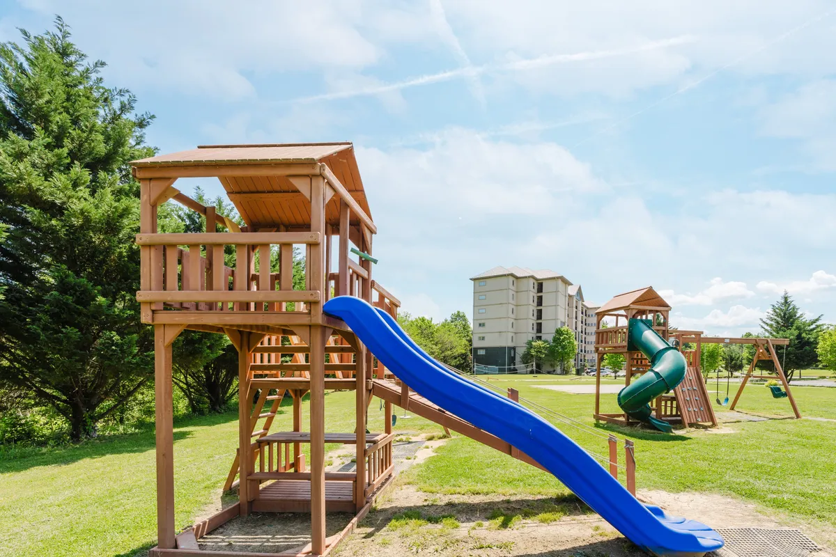 A cheerful outdoor playground scene, with a colorful structure featuring blue and green slides, surrounded by grass and open space. Children are actively playing, while families gather nearby, creating an inviting atmosphere for families seeking kid-friendly amenities.