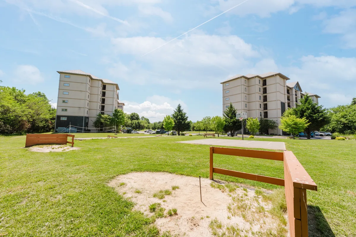 A relaxed exterior scene showing a grassy area with a basketball hoop and trees, set against the backdrop of multi-story resort buildings. The space is welcoming, designed for outdoor activities, sports, and community gatherings.