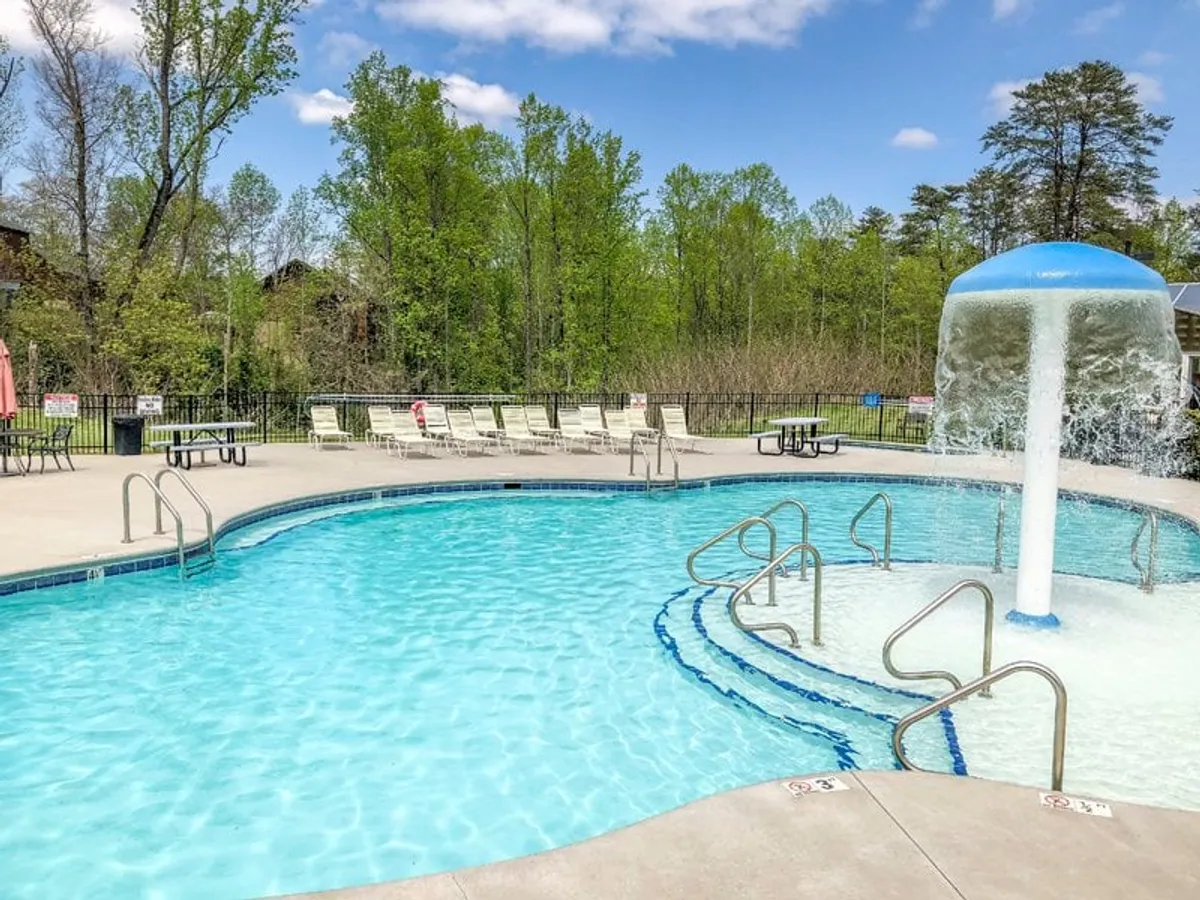 An exterior view of the outdoor pool area, featuring a sparkling swimming pool with a playful water feature, surrounded by lounge chairs and lush greenery. People are seen swimming, sunbathing, and socializing in a lively, family-friendly setting.