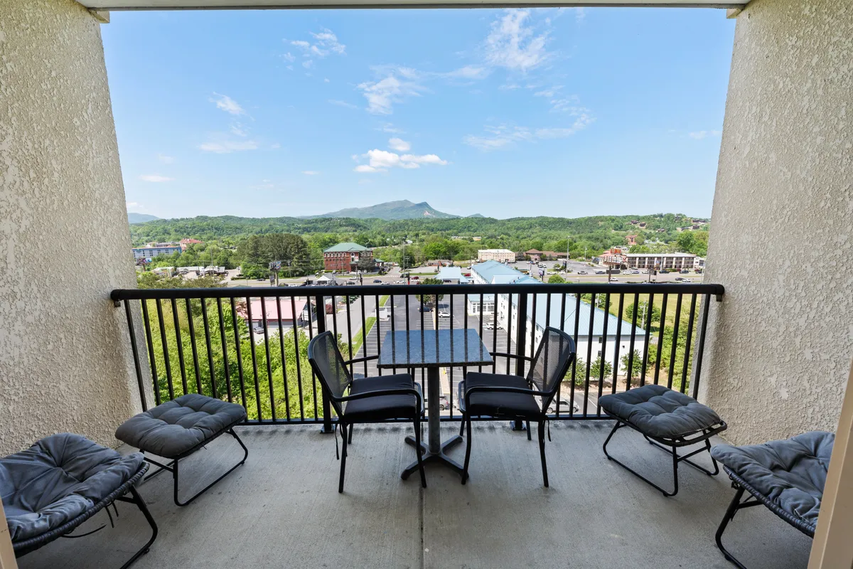 A serene balcony setup featuring comfortable outdoor furniture overlooking a vast, picturesque mountain range with the vibrant town of Pigeon Forge visible below. The sky is bright, adding to the inviting and tranquil atmosphere.