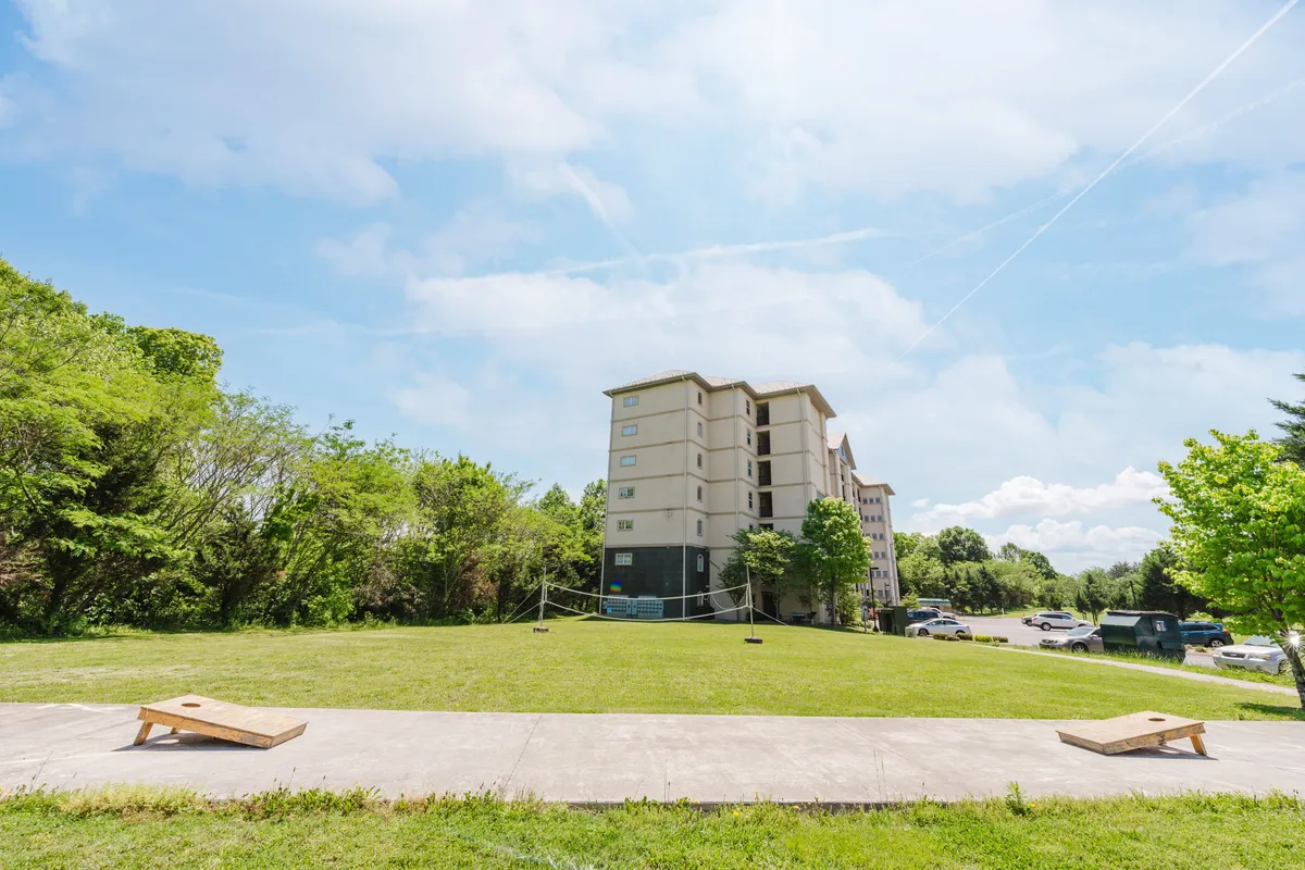 An exterior view of the multi-story building is surrounded by a green lawn with trees and cornhole boards set up, suggesting space for outdoor games and social gatherings. The relaxed atmosphere and visible amenities connect to group activities.