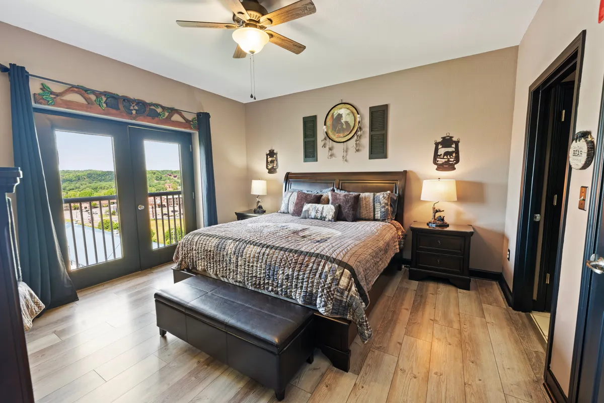 A serene master suite featuring a king-size bed dressed in plush linens, a wooden bench at the foot, large windows that frame picturesque views of mountains and the main strip, nightstands with elegant lamps, and tasteful decorative wall art. The room is bathed in warm, ambient lighting, inviting relaxation and comfort.