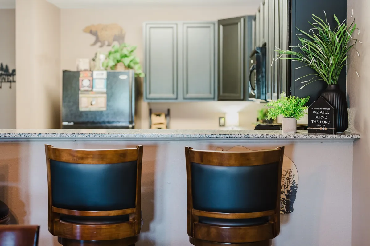 A bright, welcoming kitchen scene with bar stools at a granite countertop, stylish cabinetry, decorative plants, and a fridge adorned with magnets. The setting suggests casual dining, socializing, and the joys of both everyday routines and special celebrations.
