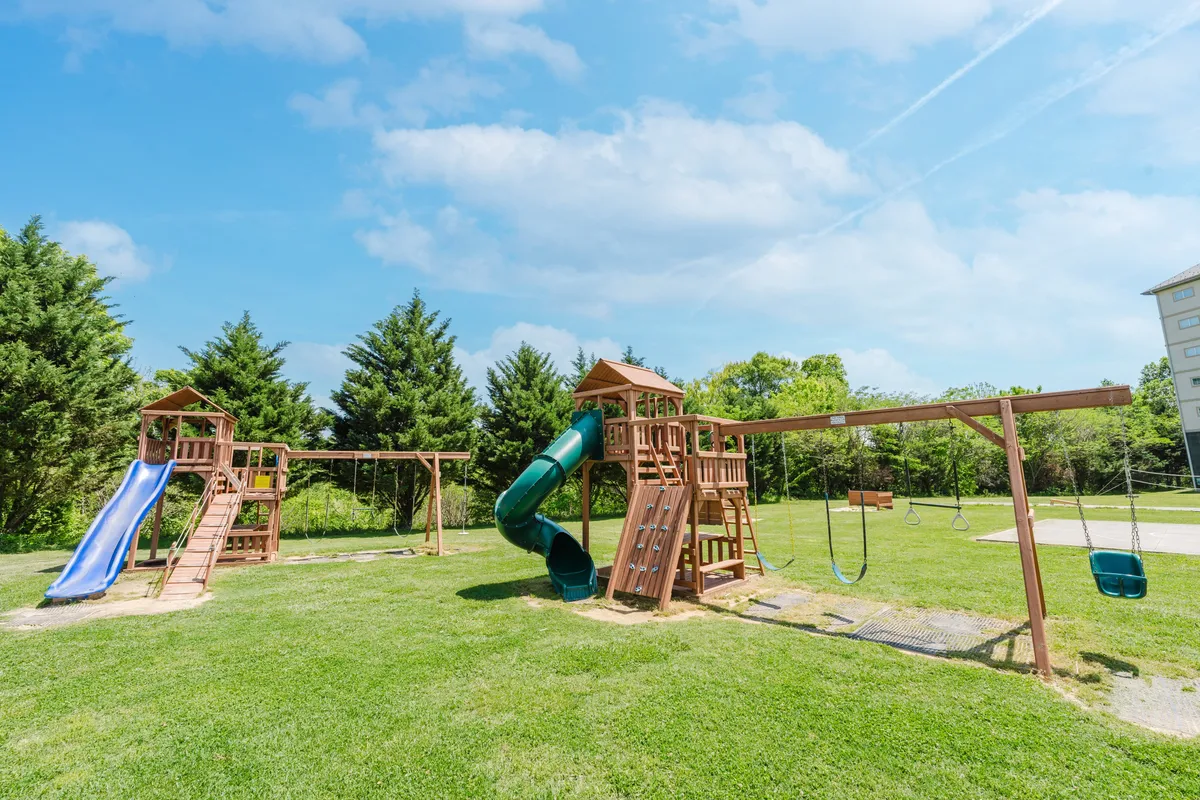 A bright and cheerful playground set in green grass, surrounded by trees. The playground equipment includes a slide and swings, with open space for children to play and families to gather. The scene radiates energy and the potential for outdoor fun.